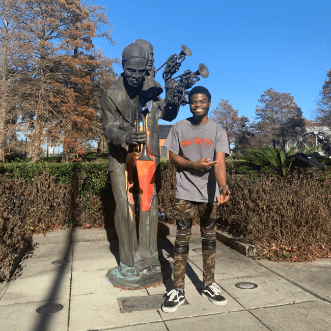 a young Black man wearing a "" shirt standing next to a Jazz statue